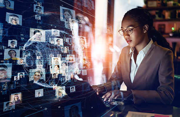Business woman of color typing on a laptop with a hologram of a globe and connections to many headshots in the foreground