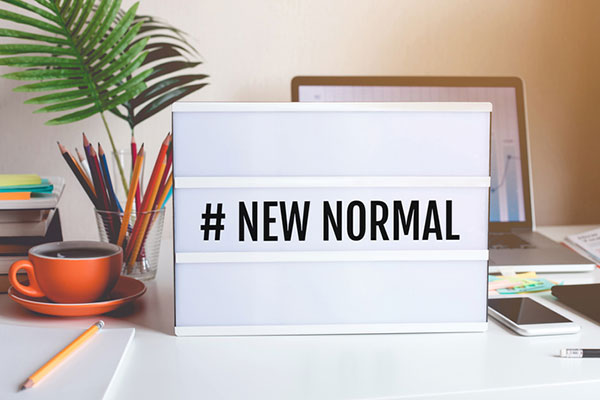 stock photo: “new normal” sign in the middle of a work desk with a cup of colored pencils, a plant, a laptop, and a cell phone