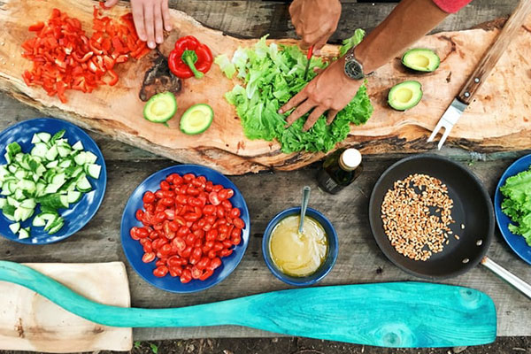 Components of a meal in preparation: cut up vegetables in bowls and on a cutting board along with a bowl of grains and of beans and a sauce. Also shows a person’s hands cutting up a green leafy vegetable