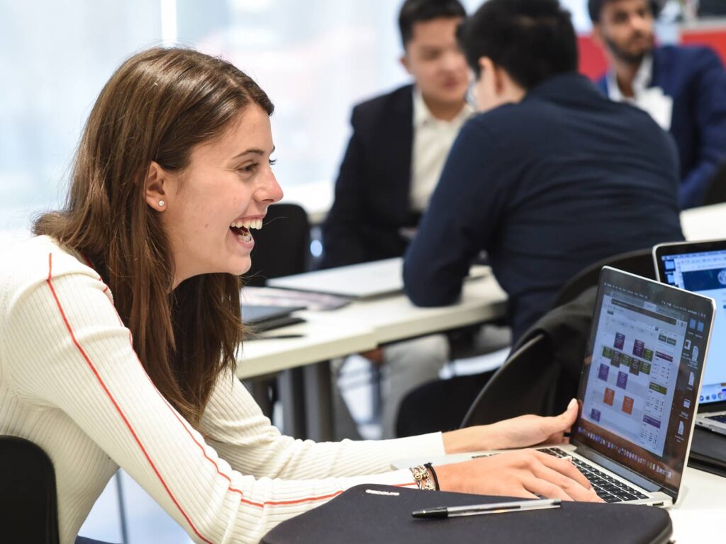 Woman smiling and laughing to someone out of frame while sitting at a table with a laptop in front of her.