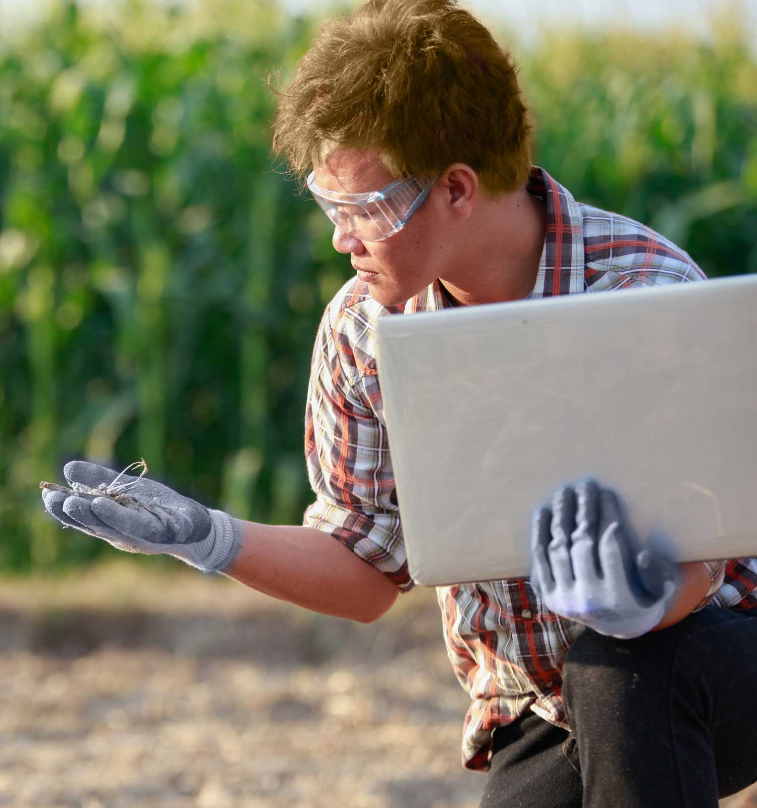 Student outside in work gloves and safety glasses holding a laptop in one hand and examining a plant in the other.