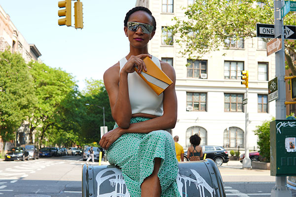 A young Black woman sitting in sunglasses, sleeveless top, and a skirt sitting on a mailbox on a city street, holding a yellow and white handbag.