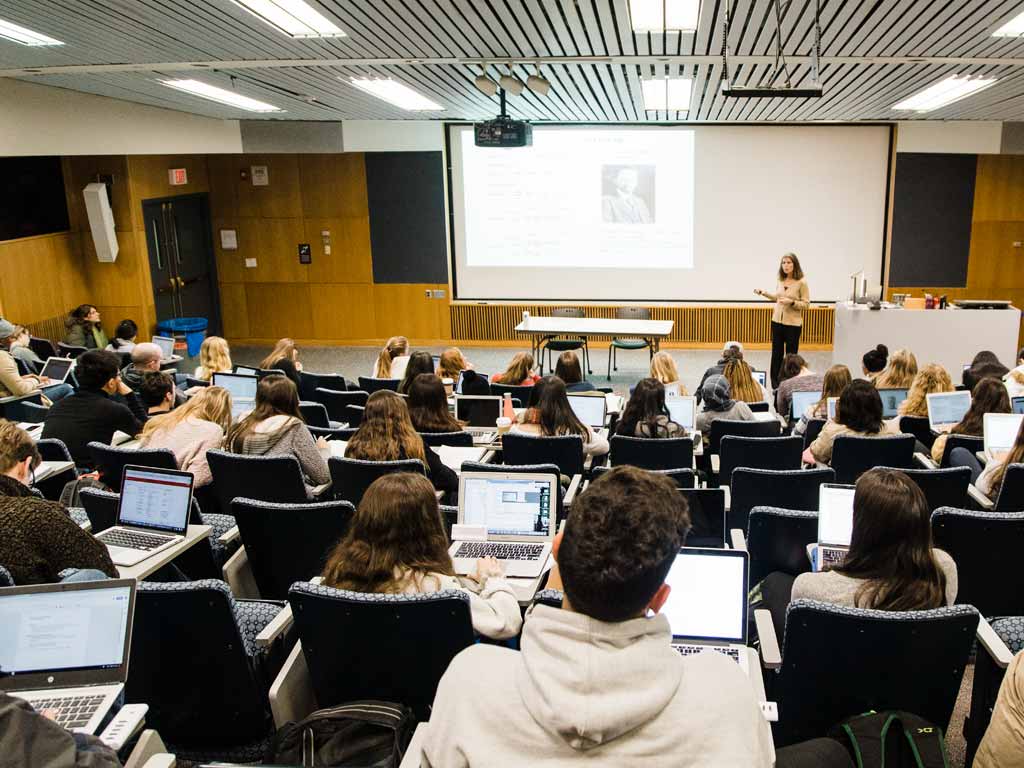 Professor stands at the front of a tiered classroom with students sitting with their laptops open in the foreground.