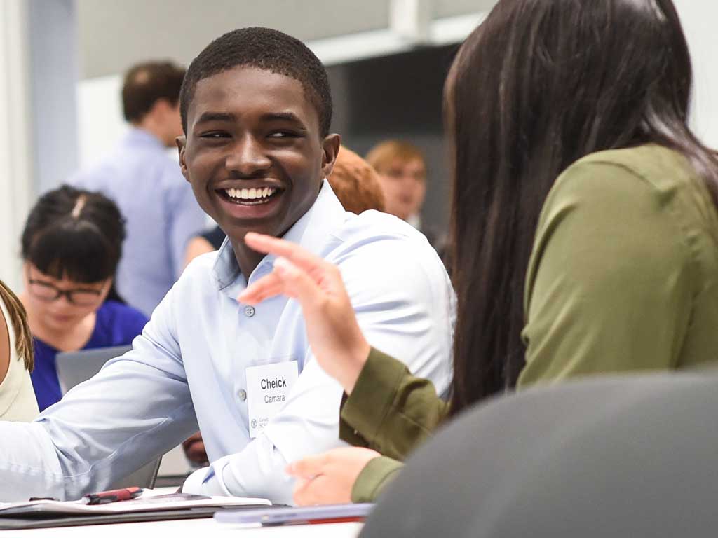 A student, in the background, smiles, and talks with another student, in the foreground.