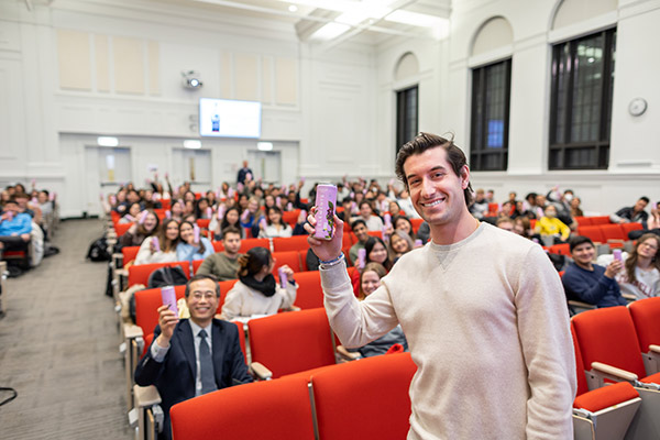 Steven Izen standing in front of a 2/3 full lecture hall, holding up a can and smiling. Many audience members are holding up similar looking cans.