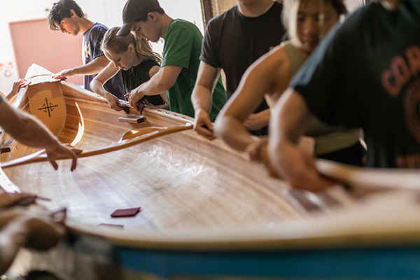 a group of students deeply focused as the work on sanding a nearly completed wooden canoe.