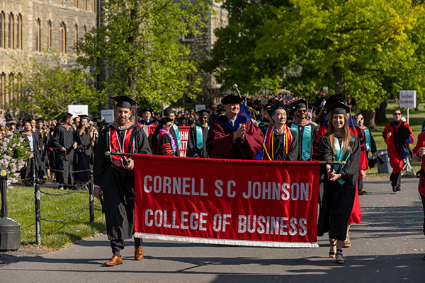 Students and faculty in caps and gowns holding and walking behind a Cornell SC Johnson College of Business banner.