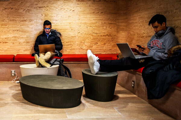 Two students sit on a bench seat on their laptops with their feet propped on tables.