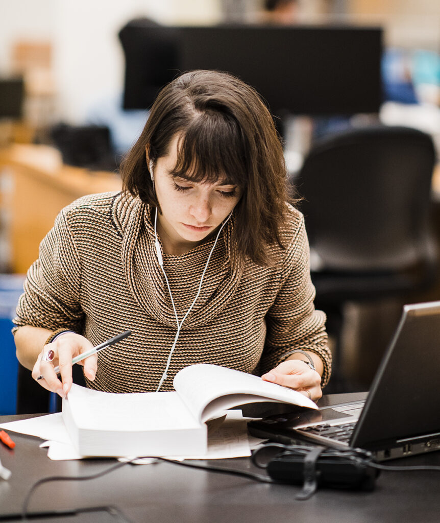 A woman wearing headphones reads a textbook with a laptop open in front of her.