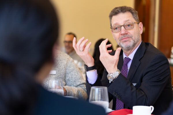 Man in a suit sitting at a table talking with both of his hands.
