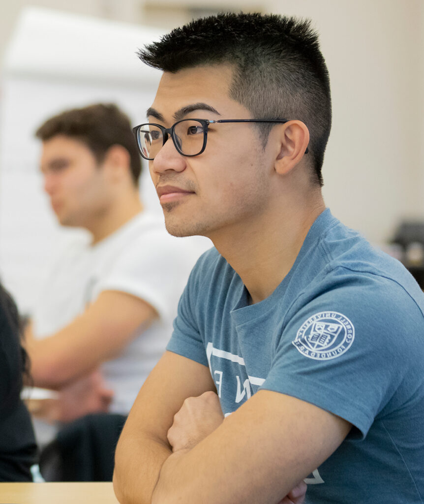 A student sits in a classroom with the Cornell University seal on his t-shirt sleeve.