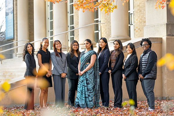 9 young women, most dressed in business attire and one in a sari, standing side by side outside a stone building in the fall.