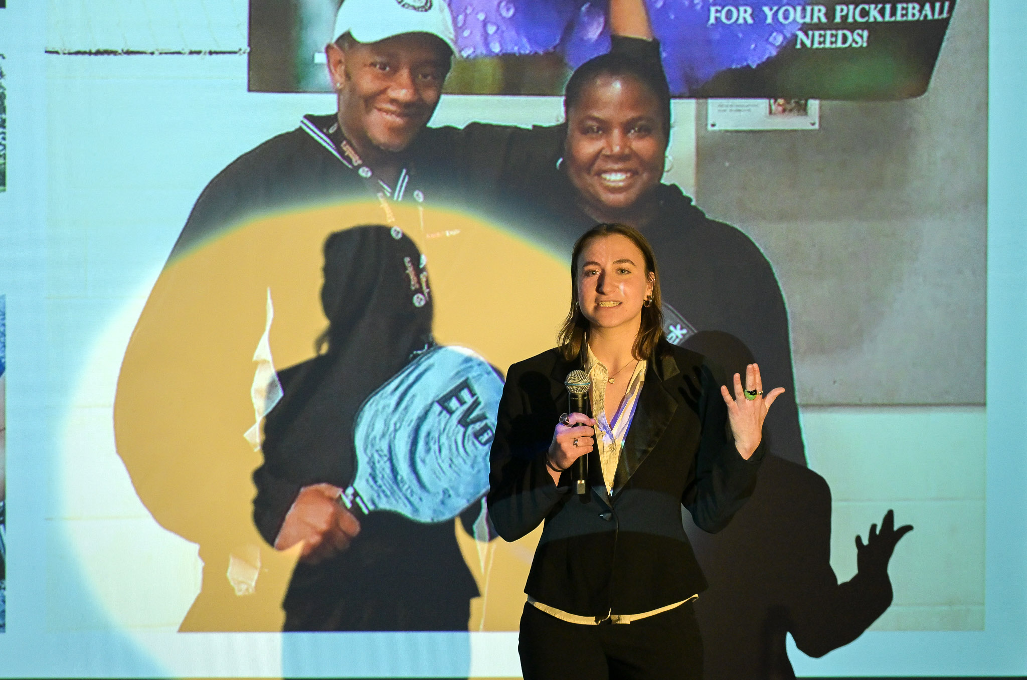 Female student speaks into a microphone in front of a screen with a photo of pickleball players.