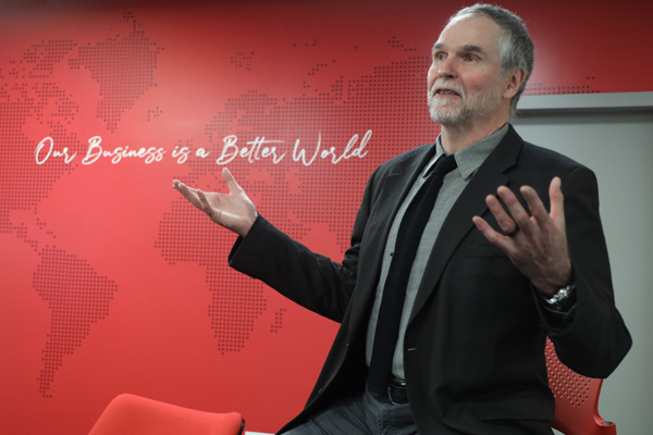 Professor John Doris sits on a stool with arms outstretched in front of a red wall with the text "Our Business is a Better World."