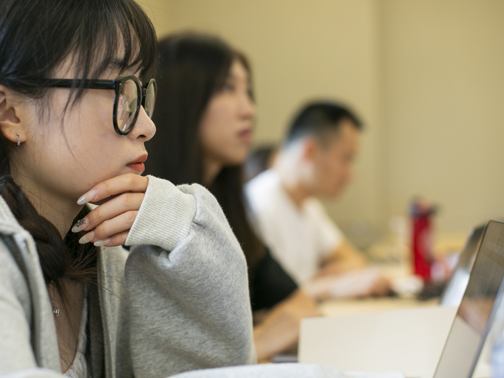 A woman sits in front of a laptop with two students visible in the background.