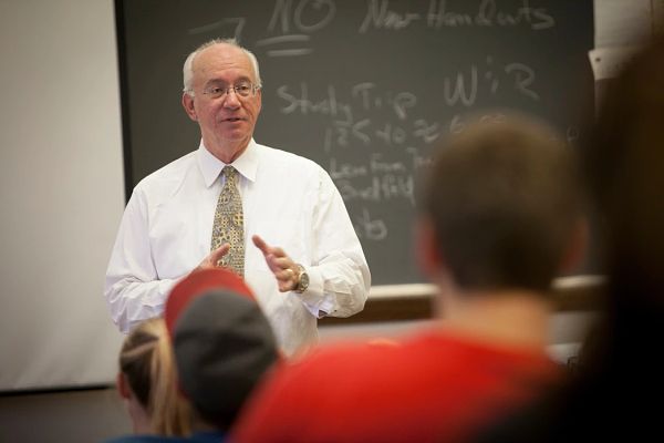 WAyne Knoblauch stands in front of a chalkboard in a white shirt and tie facing students seated in class.