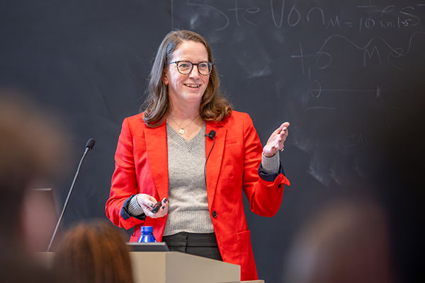 A woman standing at a podium in front of a classroom of students, speaking, smiling, and gesturing with one hand.