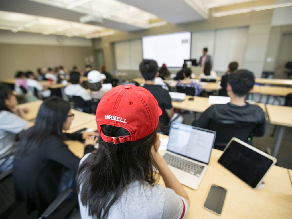 Student in a Cornell baseball hat sitting in a classroom listening to a lecture.