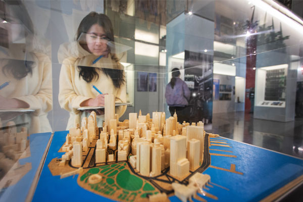 Female student takes notes while observing a wooden architectural model of a city displayed in a glass case. The background features an exhibition space with displays, informational panels.