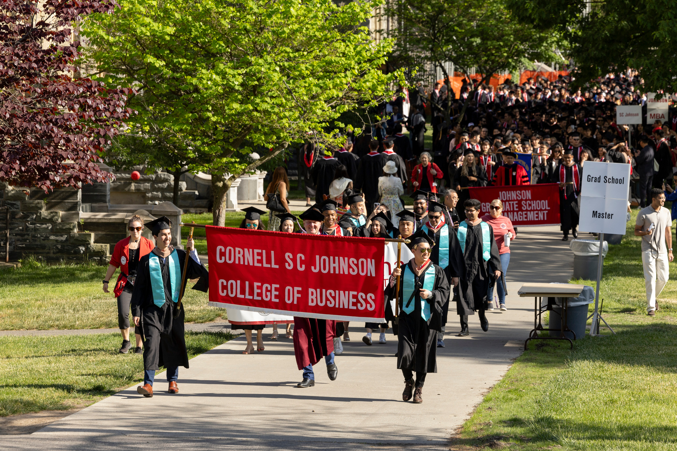 Commencement processional of students in the SC Johnson College of Business dressed in graduation regalia and holding a college banner.